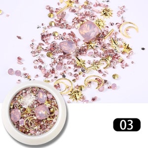 May include: A collection of nail art supplies, including pink and gold rhinestones, gems, and charms. The assortment features various shapes, such as stars, moons, and teardrops, with a small container filled with similar items. The number "03" is displayed in a black box.