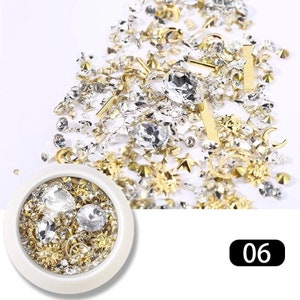 May include: A collection of nail art embellishments in a variety of shapes and sizes. The assortment includes clear rhinestones, gold stars, bars, and floral designs. A small white container holds a selection of the same items. The number "06" is in the bottom right corner.