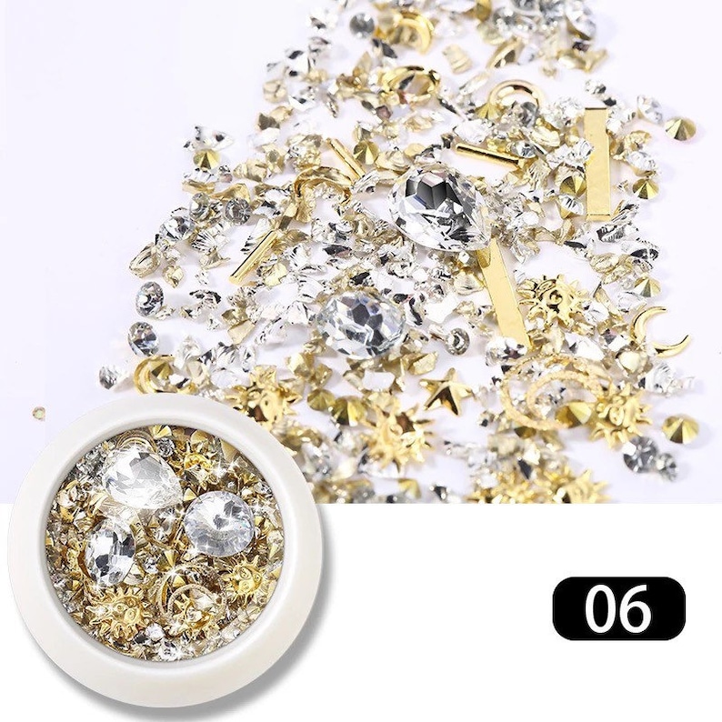 May include: A collection of nail art embellishments in a variety of shapes and sizes. The assortment includes clear rhinestones, gold stars, bars, and floral designs. A small white container holds a selection of the same items. The number "06" is in the bottom right corner.