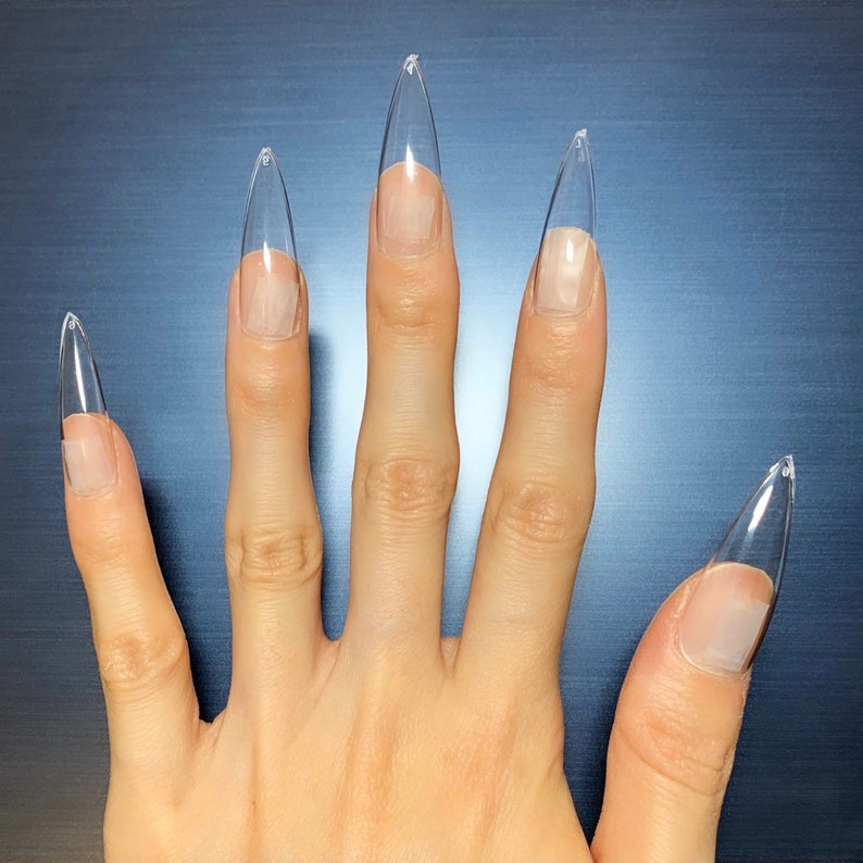 May include: A hand with five clear, stiletto-shaped acrylic nails. The nails are long and pointed, extending beyond the natural nail bed. The background is a dark, solid color.
