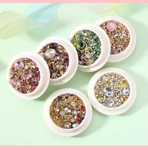 May include: Six white containers filled with various nail art embellishments. The containers hold a mix of rhinestones, gems, and metallic charms in colors like pink, gold, green, and clear. The arrangement is on a light green surface.