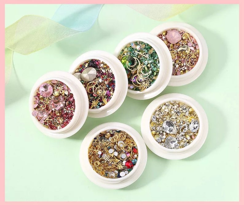 May include: Six white containers filled with various nail art embellishments. The containers hold a mix of rhinestones, gems, and metallic charms in colors like pink, gold, green, and clear. The arrangement is on a light green surface.