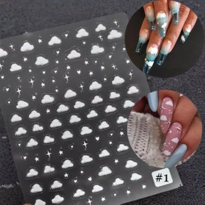May include: Clear nail art stickers featuring a pattern of white clouds, stars, and hearts. The stickers are on a transparent sheet. Two examples of nail art using the stickers are shown, one with long, clear nails and the other with pink and blue nails.