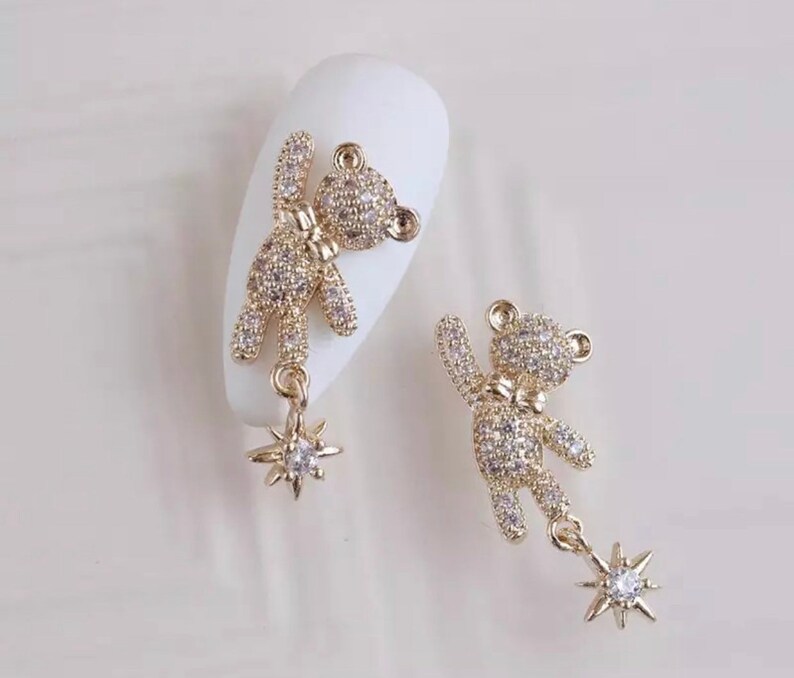 K&ouml;nnte beinhalten: Zwei goldfarbene B&auml;ren-Charms, besetzt mit klaren Kristallen. Jeder B&auml;r hat eine Fliege und einen baumelnden sternf&ouml;rmigen Charm mit einem einzelnen Kristall. Die Charms sind f&uuml;r Nailart konzipiert.