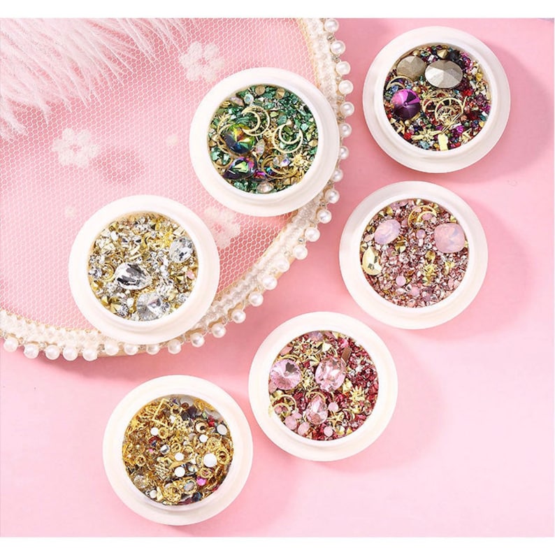 May include: Six small, white, round containers filled with various nail art embellishments. The containers hold a mix of rhinestones, gems, and metallic charms in gold, silver, and iridescent colors. The background is a pink surface with a lace doily.
