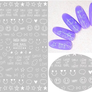 May include: A sheet of nail art stickers with white drawings on a gray background. Designs include stars, smiley faces, clouds, hearts, and text like "Hello," "Smile," and "Love." Also, five purple nail tips with white designs.