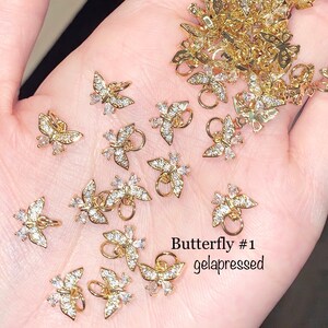 May include: Gold-colored butterfly charms with clear crystal accents. Each charm has a small loop for attachment. The text "Butterfly #1 gelapressed" is visible in the image. These charms are suitable for jewelry making or craft projects.