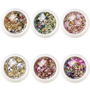 May include: Six white circular containers filled with various nail art embellishments. Each container holds a different mix of rhinestones, gold-colored metal charms, and iridescent glitter in shades of gold, silver, pink, green, and purple.