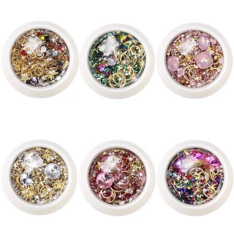 May include: Six white circular containers filled with various nail art embellishments. Each container holds a different mix of rhinestones, gold-colored metal charms, and iridescent glitter in shades of gold, silver, pink, green, and purple.