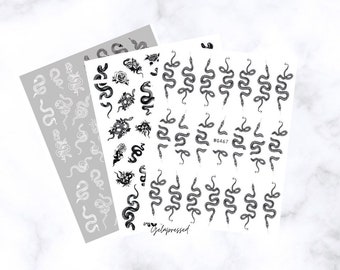 May include: Three sheets of nail art stickers featuring snake and floral designs. One sheet is light gray with white snake illustrations. The other two sheets have black snake and floral designs on a white background. The text "Gelapressed" is visible.