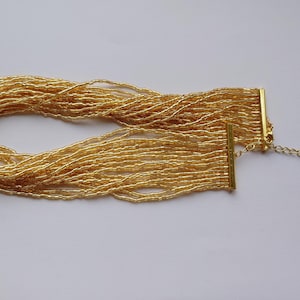 Può includere: Una collana di perline color oro con più fili. La collana ha una chiusura color oro e un'estensione a catena. Le perline, piccole e uniformi, creano un aspetto fluido ed elegante. Il gioiello è esposto su una superficie bianca.