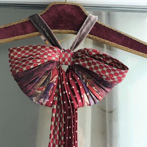 Può includere: Un papillon decorativo nei toni del rosso, bordeaux e bianco, con un motivo. Il papillon è appeso a un appendiabiti bordeaux e dorato con collo in velluto. Il papillon ha un design a pieghe e una lunga coda sfrangiata.