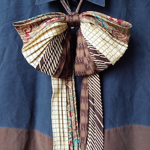 Può includere: Un papillon unico realizzato con cravatte riciclate, con una varietà di motivi e colori, tra cui marrone, bianco e blu. Il papillon è attaccato a una camicia con colletto blu e marrone.