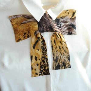 May include: A white shirt with a contrasting bow tie. The bow tie has a pattern of animal prints in brown, tan, and black. The shirt is buttoned at the top and is hanging on a white hanger.