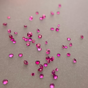 Può includere: Una collezione di numerose piccole pietre preziose rotonde e sfaccettate in un vibrante colore rosa magenta. Queste pietre sciolte a taglio brillante sono sparse nell'inquadratura, mostrando superfici riflettenti e forme circolari uniformi.