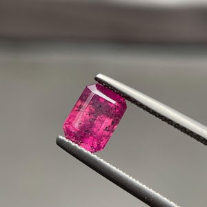 Op de afbeelding: Een levendige, rechthoekige edelsteen in smaragdgeslepen vorm met een diepe magenta-roze tint. De steen heeft donkere interne insluitingen en facetten die het licht vangen, vastgehouden door een metalen pincet met gekartelde randen.