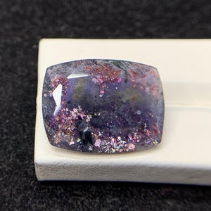 May include: A rectangular gemstone with a faceted surface, displaying a deep purple colour with pink and iridescent inclusions. The gem is set on a white surface against a dark background, highlighting its unique texture.