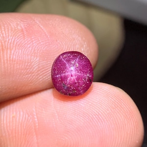 May include: A polished, oval-shaped ruby gemstone. The deep red gem features a six-rayed star pattern across its surface. The stone is held between fingers, showcasing its color and clarity. The ruby is a natural gemstone.
