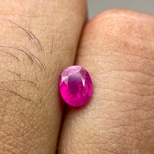May include: An oval-cut, vibrant pinkish-red gemstone with a faceted surface that reflects light. The stone displays a translucent quality and a polished finish, showcasing a brilliant saturation of color throughout its entire oval-shaped structure.