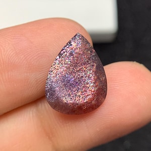 May include: A pear-shaped gemstone with a sparkling, iridescent surface. The stone exhibits a mix of colours, including purple, pink, and red, with a speckled appearance. The gem is held between fingers, highlighting its polished, smooth texture.
