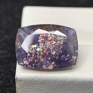 May include: A rectangular gemstone with multiple facets, displaying a deep purple colour. The gem is filled with iridescent inclusions, sparkling with pink, green, and gold. The stone is set against a white surface.