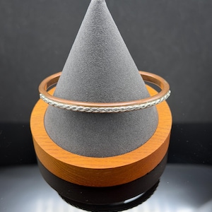 May include: A copper and silver-tone bangle bracelet. The bracelet has a copper band with a silver-tone braided design around the center. The bracelet is displayed on a gray cone-shaped stand with a wooden base.