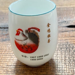 May include: White ceramic teacup with a rooster illustration and red and gold accents. The text on the cup reads "Year of: 1957 1969 1981 1993 2005".