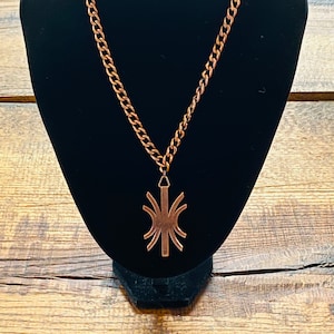 May include: A copper necklace with a geometric pendant. The pendant is a stylized design with a central vertical line and radiating lines.