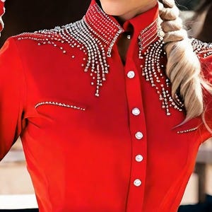 May include: A vibrant red button-down shirt with a high collar and long sleeves. The collar and shoulders are embellished with sparkling silver rhinestones. The shirt has white buttons and a tailored fit, suitable for equestrian or western wear.