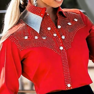 May include: A bright red Western-style shirt with intricate beadwork and silver accents. The shirt features a pointed collar with a silver metallic finish and decorative embellishments around the yoke and button placket. The long-sleeved shirt has large, round, white buttons.