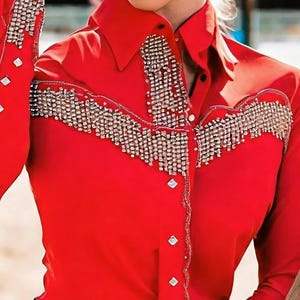 May include: A bright red button-down shirt with long sleeves. The shirt features elaborate silver rhinestone embellishments across the chest and on the sleeves. The collar is pointed, and the buttons are silver.