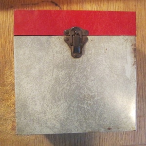 May include: A rectangular box with a red top and a light gray body. The box has a metal clasp in the center. The box is sitting on a wooden surface.