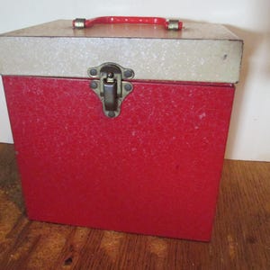 May include: A square storage case with a bright red body and a speckled off-white lid. The case features a red plastic handle on top and a central metal latch mechanism on the front. The exterior surface has a textured, speckled finish throughout.
