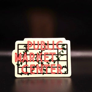 May include: A white sticker with a black grid pattern and green vines. The text "PUBLIC MARKET CENTER" is in red neon lettering.