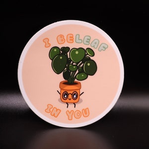 May include: A round sticker with a cartoon plant in a terracotta pot. The plant has large green leaves. The sticker has the text "I BELEAF IN YOU" in orange letters. The background is a light peach color with a white border.