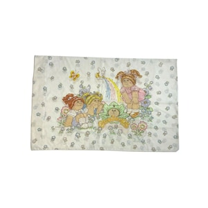 Pode incluir: Uma fronha branca com um padrão floral e uma estampa de três bonecas Cabbage Patch Kids. As bonecas estão sentadas em uma flor verde com um arco-íris atrás delas. O texto "Cabbage Patch Kids" está impresso na fronha.