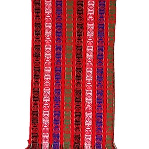 Op de afbeelding: Een rechthoekig geweven textiel met een levendige rode achtergrond en verticale strepen. De strepen hebben geometrische patronen in wit, blauw, groen en zwart. De onderkant heeft een franje. Dit textiel is waarschijnlijk een sjaal.