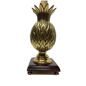 Puede incluir: Lámpara de mesa de latón con forma de piña y base de madera oscura. La lámpara presenta texturas detalladas en forma de hojas y un tono dorado cálido. La base es cuadrada, con pequeños pies redondos.