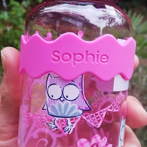 Könnte beinhalten: Rosa Plastikwasserflasche mit einem rosa Silikonband, auf dem "Sophie" steht. Die Flasche hat ein rosa-weißes Karomuster und ein Cartoon-Eulen-Design.