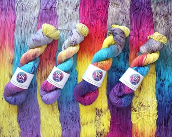 May include: Four skeins of vibrant, multi-coloured yarn in a rainbow gradient. The colours include yellow, blue, purple, and pink. Each skein has a white label with the text "VK Yarns". The background is made of vertical yarn strands in similar hues.
