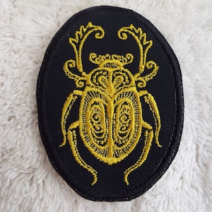 May include: A black oval patch with a gold embroidered beetle design. The beetle has intricate details and a stylized design.