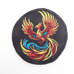 May include: Embroidered patch featuring a phoenix design. The patch is round with a black background. The phoenix is depicted in shades of red, yellow, and blue, with outstretched wings and a flowing tail. Ideal for jackets, bags, or other fabric items.