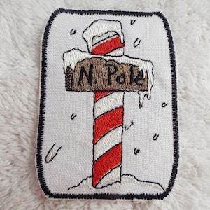 Pode incluir: Etiqueta bordada com um poste de pau de cana com uma placa que diz "N. Pole" e neve no topo. O poste é listrado de vermelho e branco e está rodeado de neve branca.