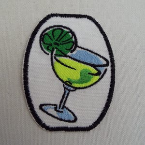 Op de afbeelding: Een witte geborduurde patch met een groene limoenwig en een blauw en geel margarita-glas.