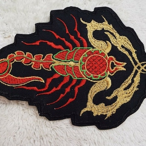 May include: A black embroidered patch with a red and gold scorpion design. The scorpion's body is red with green accents, and its tail is curled up. The scorpion is surrounded by a gold design.