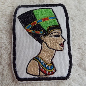 May include: Embroidered iron-on patch featuring a profile of an Egyptian queen wearing a green, black, and gold headdress. The queen is wearing a gold necklace and a red, white, and blue patterned dress.