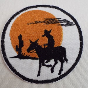 May include: Embroidered patch featuring a black silhouette of a cowboy riding a donkey against an orange sunset. The patch is circular and has a black border.