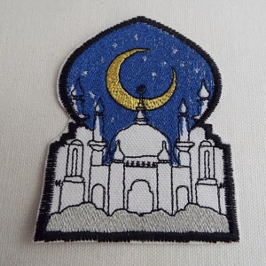 Puede incluir: Un parche bordado que representa una mezquita blanca con una luna creciente y estrellas sobre un fondo de cielo azul nocturno. La mezquita tiene múltiples minaretes y un techo abovedado.