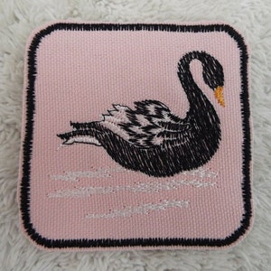 May include: Black embroidered patch of a swan swimming on pink fabric with a black border.