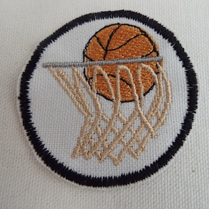 以下が含まれることがあります： バスケットボールがネットを通過している刺繍パッチ。バスケットボールはオレンジと茶色、ネットはベージュ、背景は白で黒い縁取りがあります。